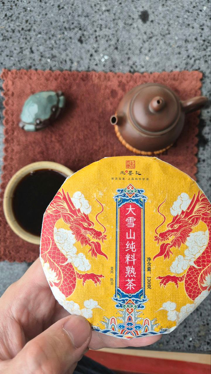 Блин шу пуэра 大雪山纯料熟茶 (Да Сюэ Шань) в руке над чайным столом: глиняный чайник, чашка тёмного чая и фигурка жабы на сером камне